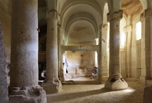 A young beautiful girl is in the pillared hall of an old, antique, underground church carved out of a sandstone cliff in Cappadocia