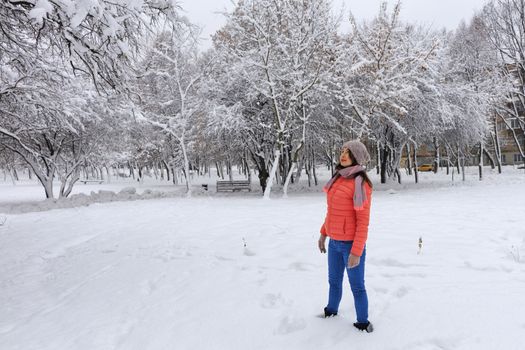 Shot of a happy and stylish beautiful woman in a bright coral jacket and blue jeans, smiling, turning her body and head, dreamily looks in the upper left corner of the snow-covered tree enjoying the winter day on the left side of copy space against the background of a winter park.
