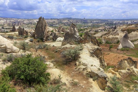 A wild rocky landscape among conic rocks, ancient caves, burnt grass and thorny shrubs runs a winding path against the backdrop of the mountainous landscape of the valley in Cappadocia.