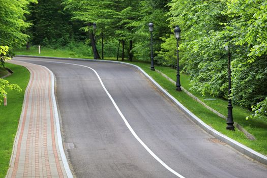 The winding asphalt road goes down the slope through the green summer park, the pavement for pedestrians is paved with stone, a number of beautiful vintage lamps on the side of the road.