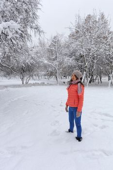 Shot of a happy and stylish beautiful woman in a bright coral jacket and blue jeans, smiling, turning her body and head, dreamily looks in the upper left corner of the snow-covered tree enjoying the winter day on the left side of copy space against the background of a winter park.