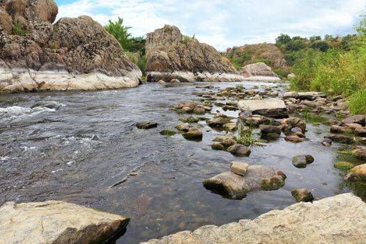 The rapid flow of the river, rocky shores, rapids, bright green vegetation and a cloudy blue sky in summer. Southern Bug River. Ukraine.