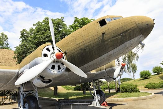 An old transport military aircraft Lisunov Li-2 of the Second World War, stands in a park against a cloudy blue sky.