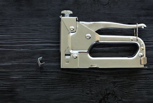 Metal industrial chrome stapler and brackets to it lie on a black wooden table with the texture of old wood.