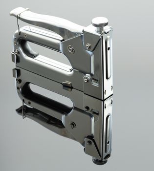 Metal industrial chrome stapler stand on the mirror surface of a gray background with a slight gradient