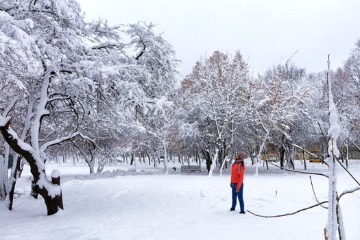 Young beautiful woman in a bright coral jacket and blue jeans in winter walks of snow-covered fairy-tale park with wooden benches