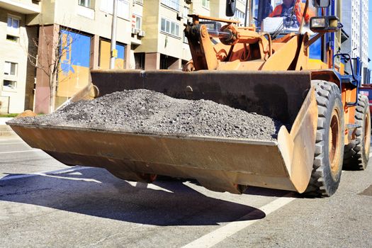 Heavy construction bulldozer carries old worn asphalt in a ladle during the construction of a road on a city street at noon