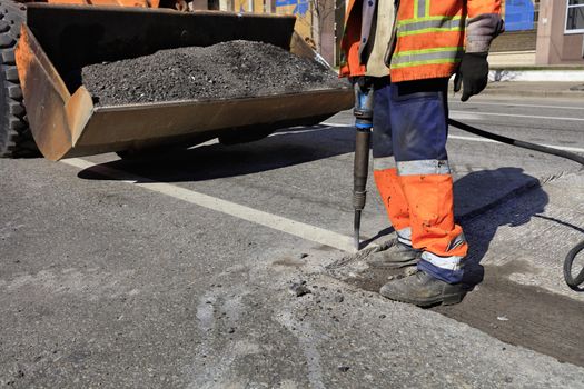 A road maintenance worker removes old asphalt on the roadway with a jackhammer into an excavator during road construction.