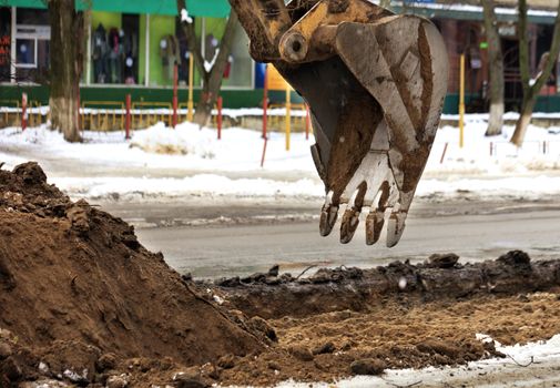 The teeth on the old excavator bucket hovered over the earthen gutter while repairing the roadway on a city street in winter.