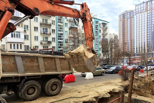 A heavy construction excavator bucket lifts sand into the back of a truck during a collector repair on a city street.