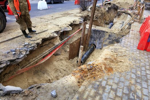 A road service worker stands near an excavated trench on the roadway and inspects concrete blocks, wells, city sewer pipes and paving slabs.