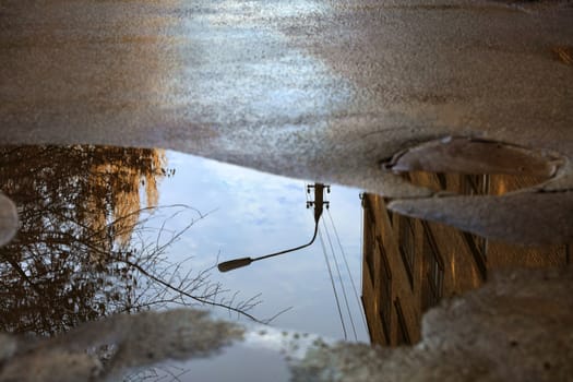 Mirror blurred reflection after a rain of the blue cloudy sky, the silhouette of a street lamp and a tree sunlit in a puddle on the asphalt.