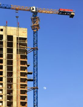 A fragment of the facade of a house under construction, a modern building is being built, an arrow of a tower crane is directed parallel to the roof against a blue cloudless sky and moon.