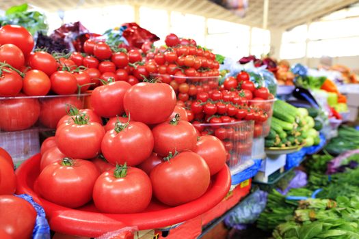Different varieties of ripe red tomatoes in the background in a blur of cucumbers, spinach and other vegetables sold in trays on the market