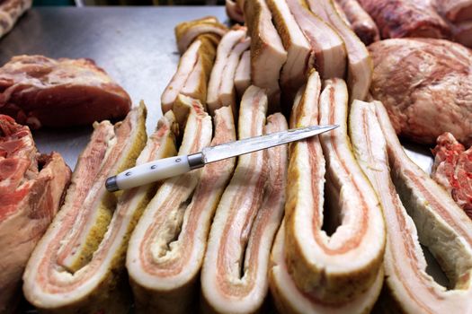 A sharp stainless steel knife, fresh salted bacon and sliced meat on the counter for sale in the market.