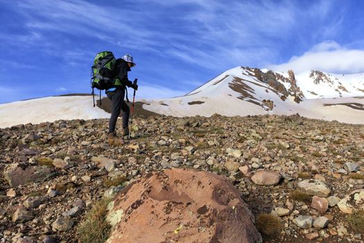 Woman tourist with a backpack on her back goes hiking on a rocky slope to a snowy mountain