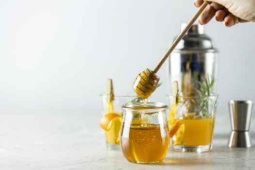 Woman hand holds a spoon for honey over jar in front of two glasses of honey bourbon cocktail with rosemary syrup or homemade whiskey sour with orange peel and rosemary decoration and bartender tools