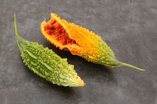 Two ripe bitter melons - split orange with red seeds, and warty green flesh - on a slate grey background 