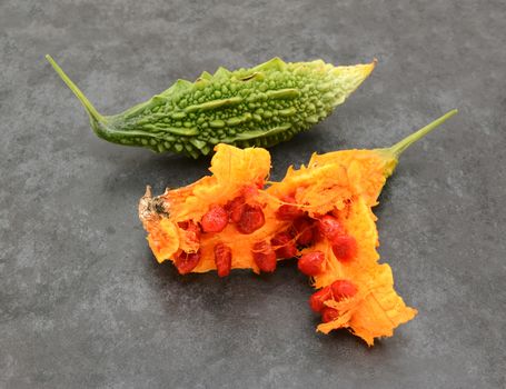 Green, ridged bitter gourd and an overripe orange fruit, split apart to show sticky red seeds - on a slate grey background