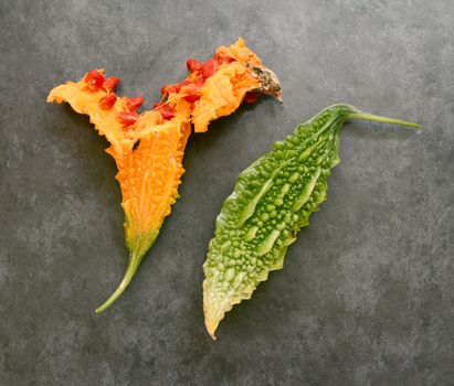 Orange, overripe bitter gourd split open with red, sticky seeds and a whole green bitter melon - on a slate gray background