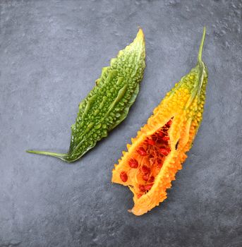 Two bitter melons - with green ridged skin and overripe orange flesh, split to show red seeds - on a slate gray background 