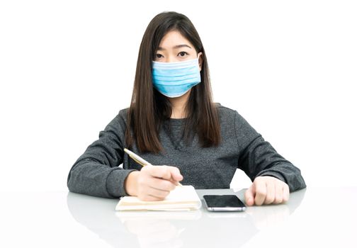 Teenage female student preparing for learnning at home sitting at a desk in front of smartphone with notepad and wear protective medical mask for protect Covid-19 or corona virus disease