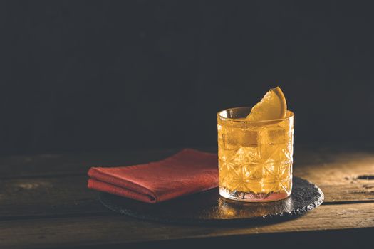 Glass of the cocktail negroni with red napkin and black stone tray on a old wooden board. Decorated orange slice. Nice romantic backlight.