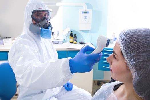 Doctor in protective suit uniform and mask checking temperature of young female patient. Coronavirus outbreak. Covid-19 concept.