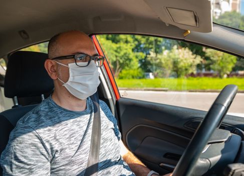 Turin, Piedmont, Italy. Maggio2020. Coronavirus pandemic: portrait of a Caucasian man driving a car wearing a white mask to avoid contagion. Selective focus on man and blurred background. Sunny day.