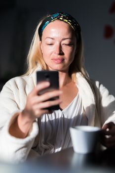 Beautiful caucasian woman at home, feeling comfortable wearing white bathrobe, taking some time to herself, drinking morning coffee and reading news on mobile phone device in the morning.