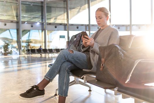 Casual blond young woman talking on cell phone while waiting to board a plane at airport departure gates. Empty airport terminal due to corona virus pandemic.