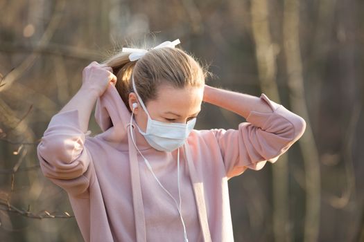 Portrait of caucasian sporty woman wearing medical protection face mask while walking in park, relaxing and listening to music. Corona virus, or Covid-19, is spreading all over the world.