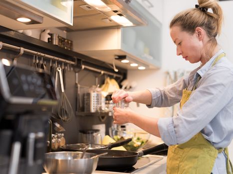 Stay at home housewife woman cooking in kitchen, salting dish in a saucepan, preparing food for family dinner. Blonde caucasian model in her 30is, real cooking not looking in camera.