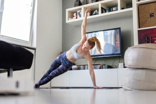 Attractive sporty woman working out at home, doing pilates exercise in front of television in her living room. Social distancing. Stay healthy and stay at home during corona virus pandemic.