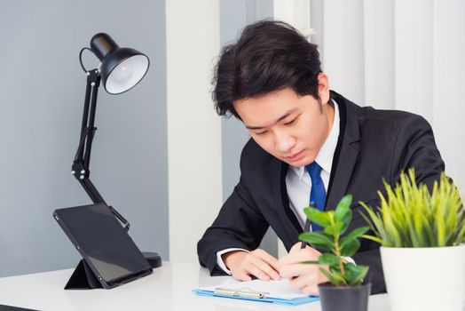 Work from home, Asian young businessman video conference call or facetime he smiling looking to paper sitting on desk using smart digital tablet computer and write notebook information at home office