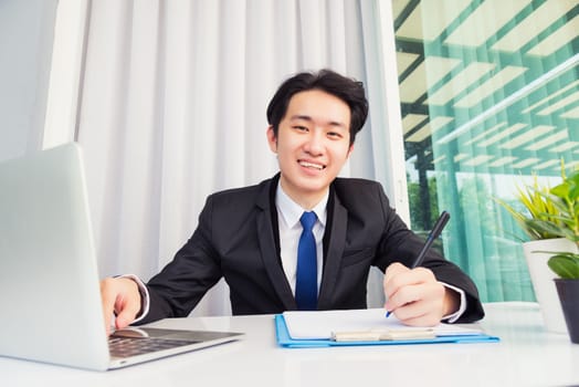 Work from home, Asian young businessman smile wearing suit video conference call or facetime by laptop computer on desk and write taking notes on paper at home office