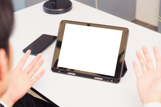 Work from home, Back view of Asian young businessman smile wearing suit video conference call or facetime by smart digital tablet computer blank screen raise his hand explain to teammates on desk