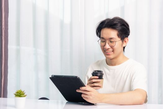 Happy Asian young business handsome man work from home office wear glasses, t-shirt comfortable he smiling and using a black modern smart digital tablet computer to read email or learning on desk