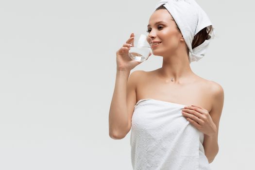 Charming pleasant woman in fluffy white towel on head with glass of clean water looking away touching towel on body