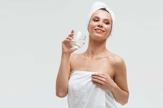 Charming pleasant woman in fluffy white towel on head with glass of clean water looking away touching towel on body