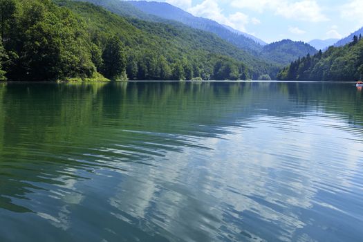 In the clear waters of a mountain lake in the deciduous forest white clouds are reflected against the blue sky