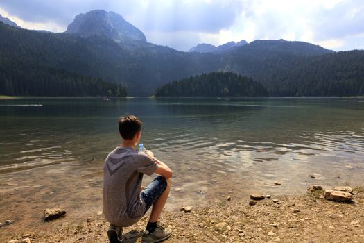 A teenager sits in the early morning on the shore of a mountain lake with a bottle of water and looks at the surface of the water.