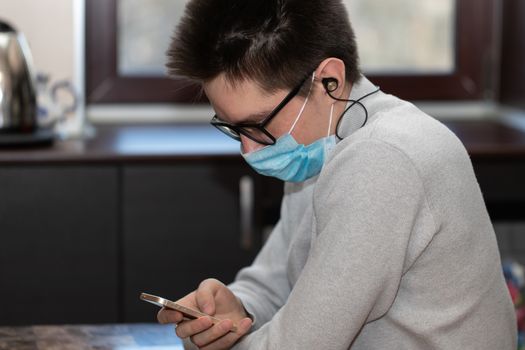 A young man in a reusable medical mask looks at the phone screen and reads messages from friends. Communication during a pandemic.