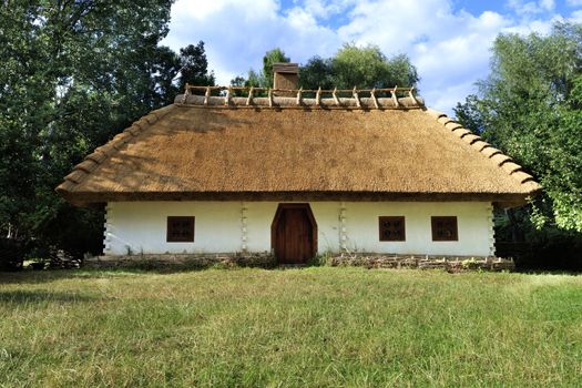 Old traditional Ukrainian rural house with a thatched roof and a wicker fence in the garden with green grass and copy space against a blue sky with a white cloud