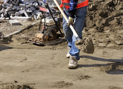 Worker aligns the foundation with sand and a shovel for laying paving slabs on the background of a gasoline rammer and a prepared area for work, close-up view.