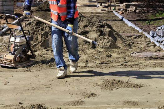 Worker aligns the foundation with sand and a shovel for laying paving slabs on the background of a gasoline rammer and a prepared area for work.