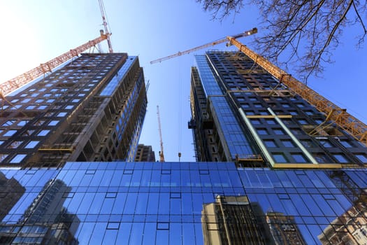 Fragment of the facade of a modern building under construction made of glass and concrete, reflected in the glass walls of adjacent buildings, tower construction cranes pointing upwards.