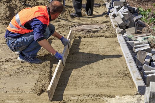 On the sidewalk, a worker cleans and levels the sandy platform with a wooden board, preparing the foundation for laying the paving slabs around the sewer manhole.