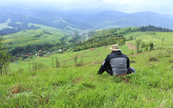 A lone traveler sits on the grass on top of a hill and looks at the picturesque landscape of the Carpathian mountains and the village in the valley, shrouded in mist.