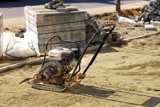 Old worn rusty gasoline compactor stands on the sand against the background of a stack of paving slabs in the blur.